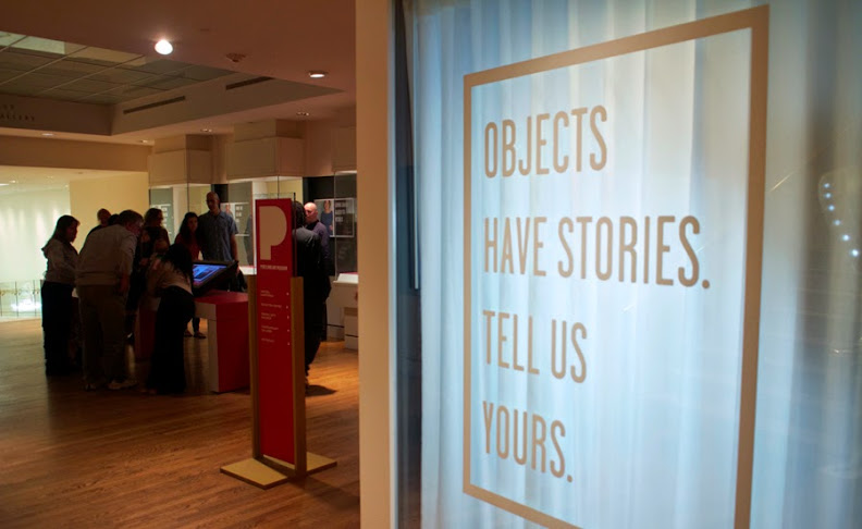 To the left of the photograph, a group of people stand around a digital touchscreen. The right of the image is a sign that says Objects have stories. Tell us yours.