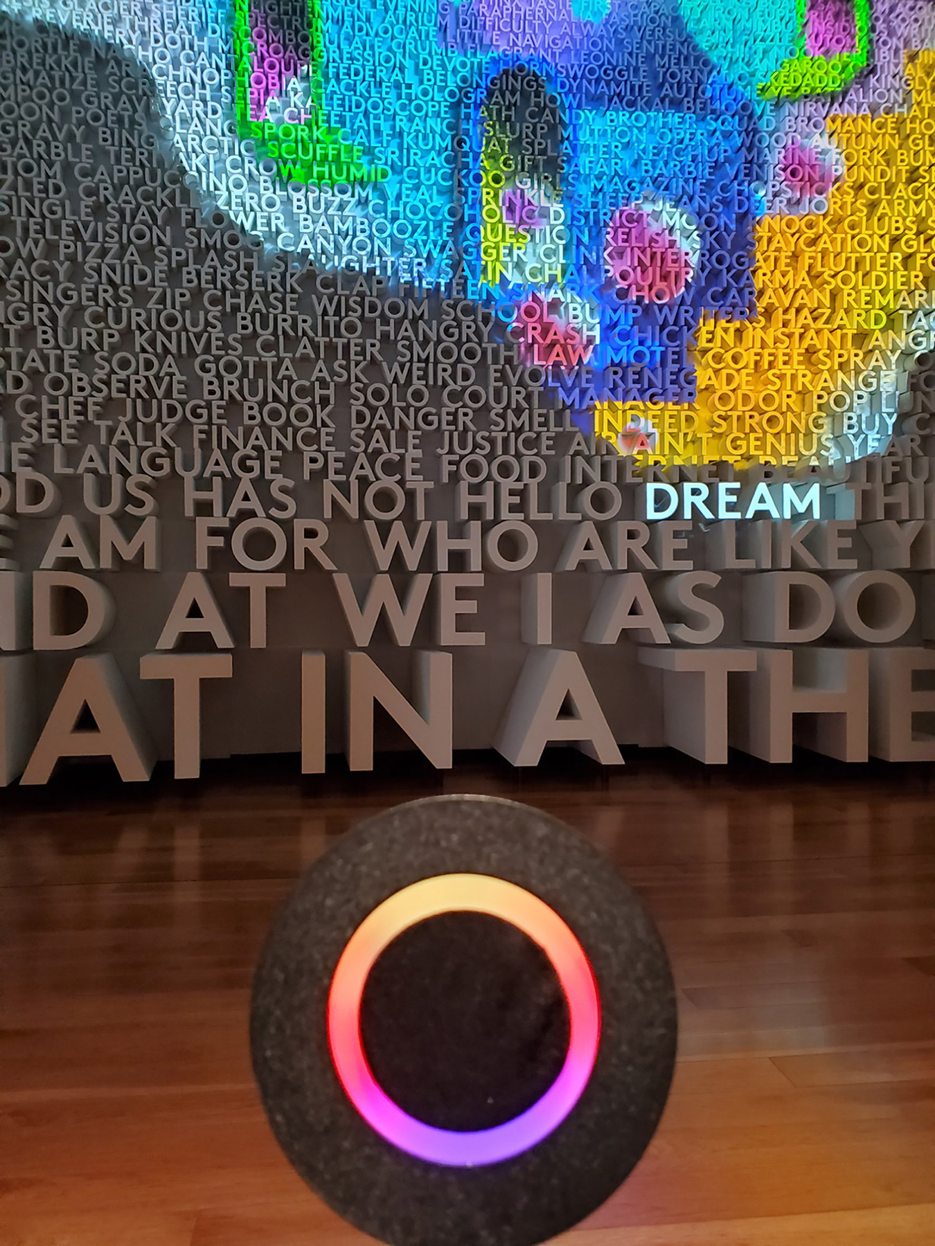 A circular microphone stands in the foreground with a rainbow light at the center. In the background three dimensional words in ascending sizes cover the wall all the way out of the frame of the photo. Projected onto the wall is a spread of abstract shapes in bright yellow, pink, blue, purple, and green. The word dream is the only purposefully highlighted word, glowing in the light from the projector.