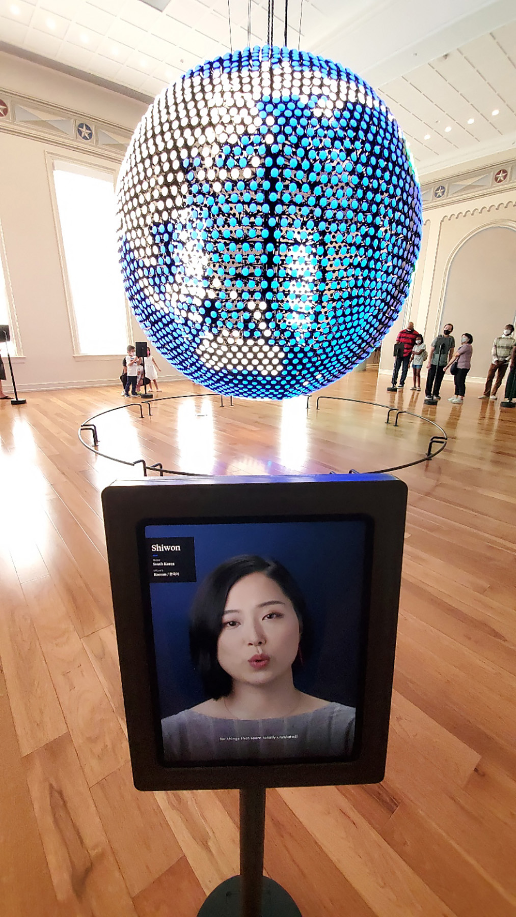 In the foreground of a bright, naturally lit room stands a touch screen with an image of a South Korean woman. She appears to be frozen mid sentence as her lips are pursed in an ooh shape. Behind the screen, suspended from the ceiling is a large globe composed of fist sized, circular LED lights which are colored to mimic the surface of the earth. This side of the globe is of Australia, Eastern Asia, and the Pacific Ocean.